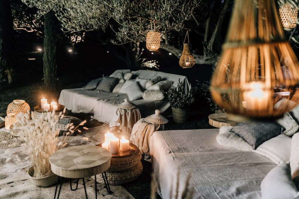 Softly lit outdoor lounge setup with hanging bulbs, white and blue pillows, and rustic wooden crates used as tables, nestled among ancient olive trees.