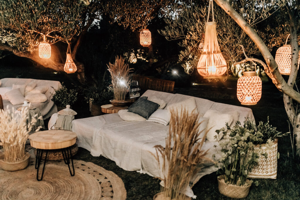 Softly lit outdoor lounge setup with hanging bulbs, white and blue pillows, and rustic wooden crates used as tables, nestled among ancient olive trees.