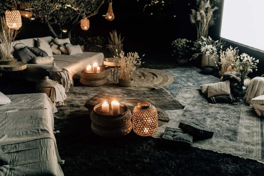 Softly lit outdoor lounge setup with hanging bulbs, white and blue pillows, and rustic wooden crates used as tables, nestled among ancient olive trees.